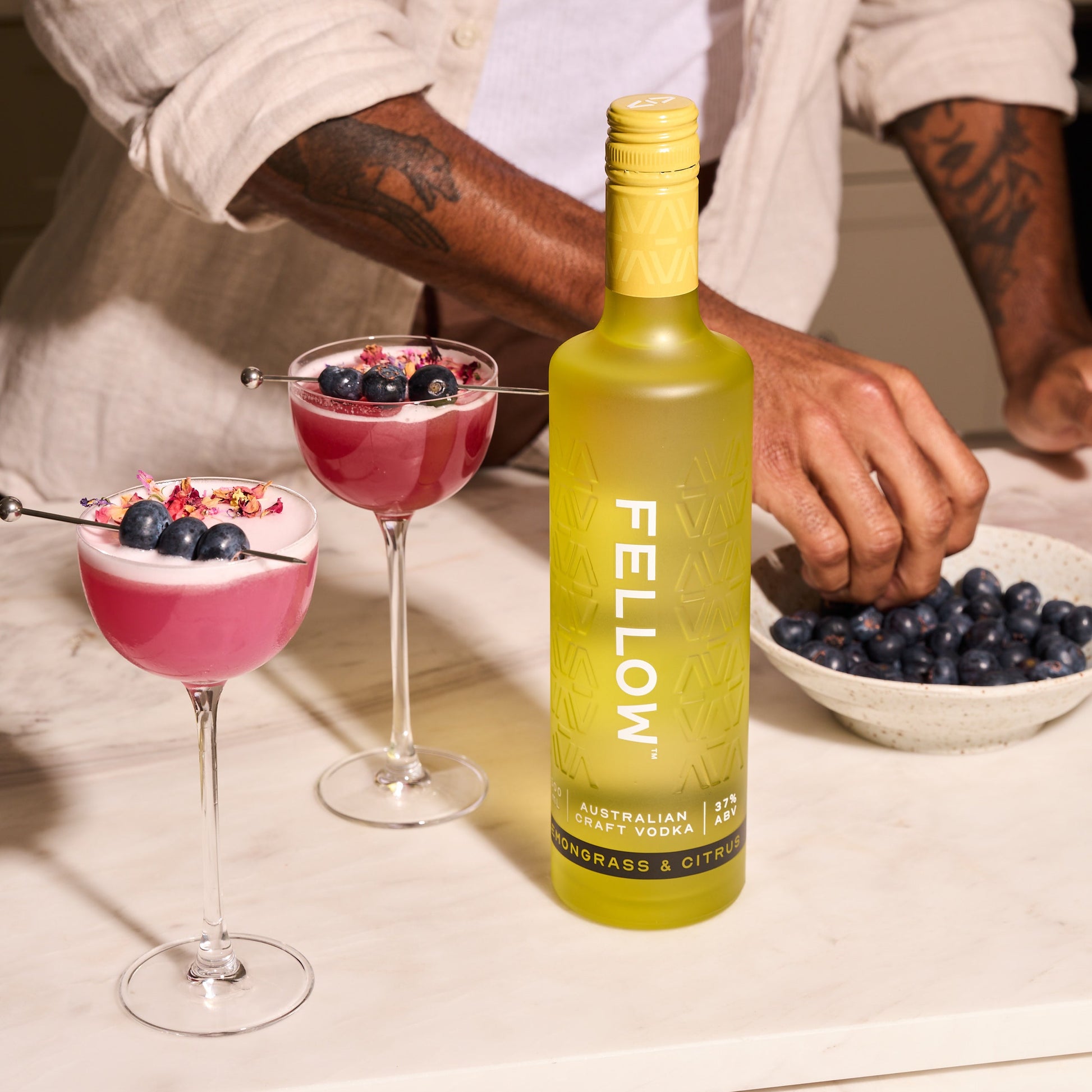 Bottle of Fellow Lemongrass & Citrus Vodka with two blueberry sour cocktails and a bowl of blueberries on a table.